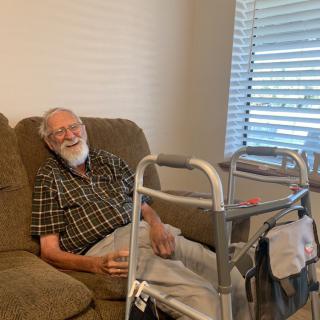 An elderly man sitting on a couch next to a walker in a living room.