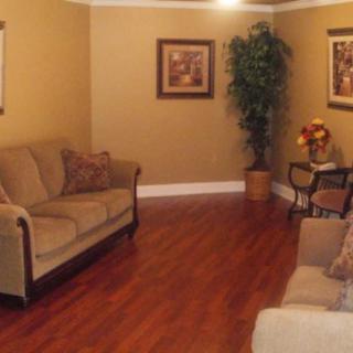 A cozy living room with wooden flooring, featuring sofas, framed artwork, and an indoor plant.