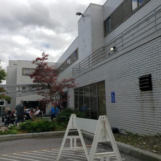 A view of a modern building with a parking area, surrounded by trees on a cloudy day.