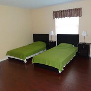 A minimalist bedroom with two twin beds, each covered in green bedding, stands on a laminate floor. A window with a valance is positioned between the beds above a nightstand.