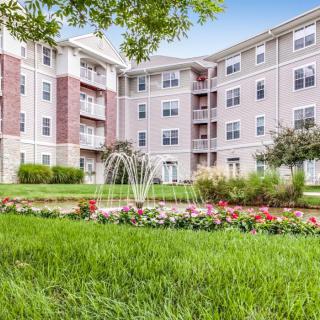 A multi-story apartment building with a landscaped garden featuring a fountain and colorful flowers.