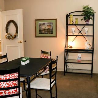 A cozy dining room with a black table and chairs, decorated with red and white cushions. A shelving unit adorned with decor items and a plant stands in the corner. The wall features artwork and a wreath on the door.