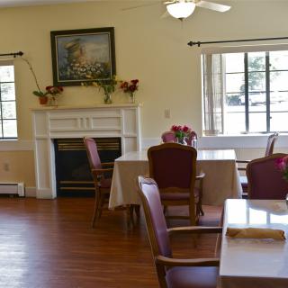 A cozy dining room with tables, chairs, and a fireplace, featuring warm decor and natural light from the windows.