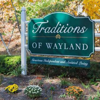 A decorative sign for Traditions of Wayland, surrounded by autumn foliage and vibrant flowers.
