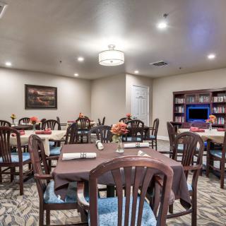 A cozy dining room with neatly arranged tables and chairs, adorned with a bookshelf and warm lighting.