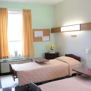 A clean and simple hospital room featuring two beds, bright curtains, and ample natural light.