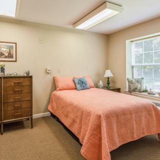 A cozy bedroom with a bed covered in a pink quilt, a dresser, and a lamp beside the window.