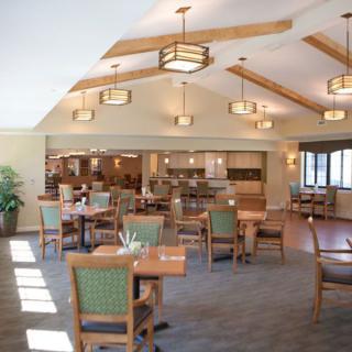 A cozy dining room with arranged tables and chairs, featuring warm lighting and wooden ceiling beams.