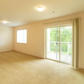 An empty apartment room with carpet flooring and a sliding glass door leading to a balcony.