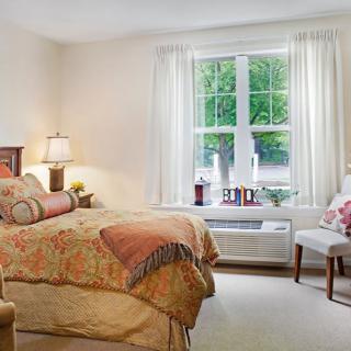 A cozy bedroom with a large window, featuring a bed with patterned bedding, a bedside lamp, and comfortable seating.