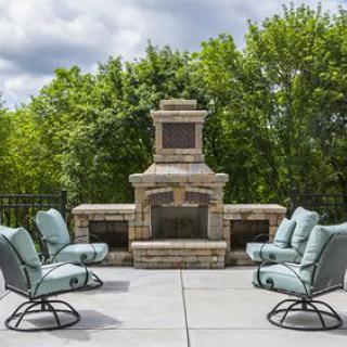 A stylish outdoor patio with a stone fireplace and cushioned chairs surrounded by lush greenery.