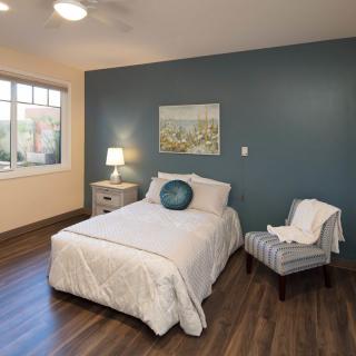 A cozy bedroom with a blue accent wall, featuring a bed, bedside lamp, chair, and large window.