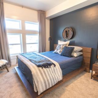 A modern bedroom with a blue accent wall and large windows, featuring a cozy bed adorned with pillows and blankets.