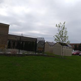 A view of a building surrounded by a fence, featuring a lone tree and grassy area under a cloudy sky.