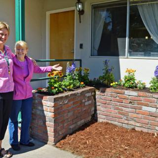 Two women proudly display a brick planter filled with colorful flowers outside a house.