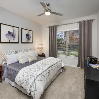 A modern bedroom with stylish decor, featuring a comfortable bed, ceiling fan, and large window with drapes.