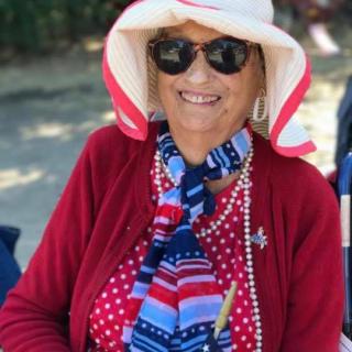 A woman wearing a stylish hat and sunglasses, accessorized with a colorful scarf, smiles outdoors.