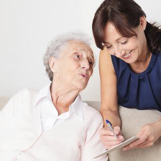 A caregiver interacts with an elderly woman, taking notes and engaging in conversation.