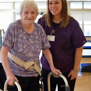 A caregiver assists an elderly woman with her walking aid, promoting rehabilitation and support in a friendly environment.