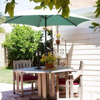 A cozy patio setup featuring a wooden table and chairs under a green umbrella, surrounded by lush greenery on a sunny day.