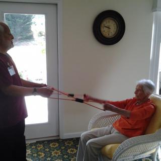 An elderly person exercising with resistance bands assisted by a caregiver in an indoor setting.