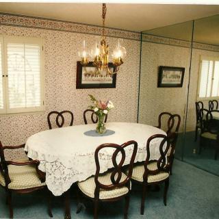A vintage dining room featuring a round table with a lace tablecloth, surrounded by ornate chairs and a chandelier overhead. A large mirror covers one wall, reflecting the floral wallpaper and framed artwork.