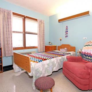 A cozy bedroom featuring a single bed with colorful bedding, an armchair, and a window with curtains.