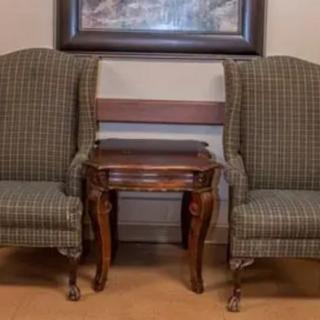 Two upholstered chairs flank a wooden side table, set against a wall with a framed painting.