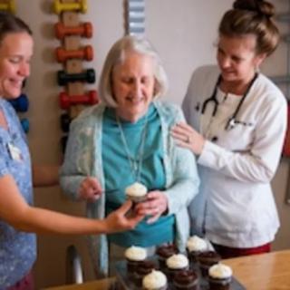 An elderly woman is joyfully celebrating with caregivers, sharing cupcakes.
