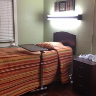 A simple room with a hospital bed covered in a striped blanket, accompanied by a side table and a bright lamp on the wall.