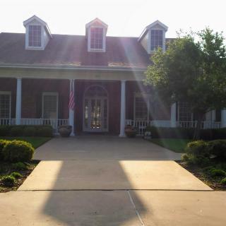 A house with a long driveway, illuminated by sunlight, surrounded by trees.