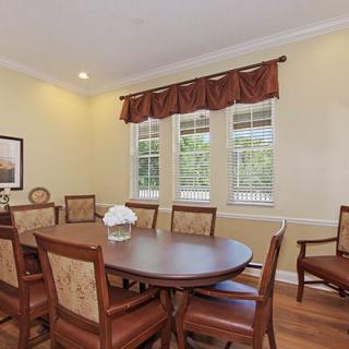 A well-lit dining room with a large table and multiple chairs, featuring elegant curtains and wooden flooring.