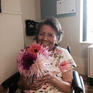 An elderly woman sitting in a wheelchair, smiling and holding a bouquet of flowers indoors.