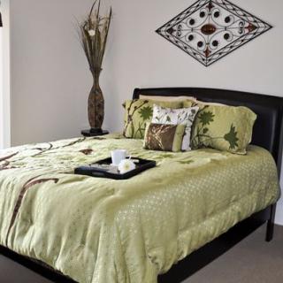 A cozy bedroom featuring a neatly made bed with green bedding and decorative pillows, complemented by wall art and a tall vase.