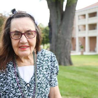 An elderly woman with glasses standing outside near a tree and building.