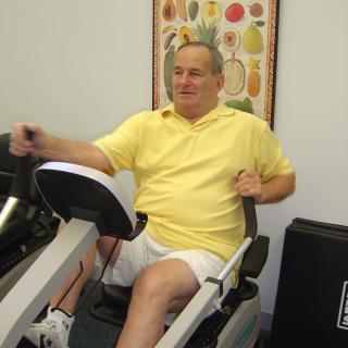 A person exercising on a recumbent bike indoors, wearing a yellow shirt and white shorts.