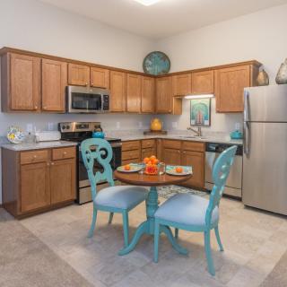 A cozy kitchen with wooden cabinets and stainless steel appliances, featuring a small dining area with blue chairs and a round table.