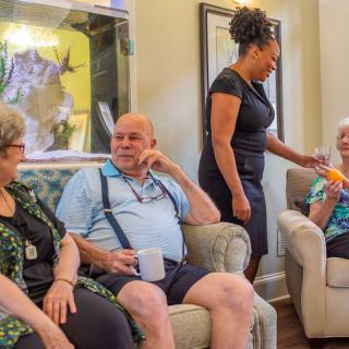 A group of seniors socializing in a living room, assisted by a caregiver.