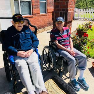 Two elderly individuals sitting in wheelchairs outside a brick building on a sunny day.