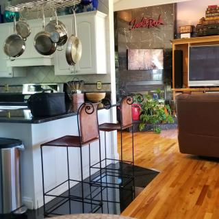 A modern kitchen with bar stools and an open living area, featuring hardwood floors and hanging pots.