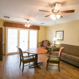 A cozy living room with a wooden floor, featuring a sofa, table, chairs, and ceiling fans.
