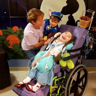 A young girl in a wheelchair enjoying time with her caregiver and a friendly costume character.