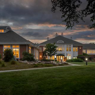 A view of a well-lit building at dusk with a cloudy sky backdrop and surrounding green landscape.