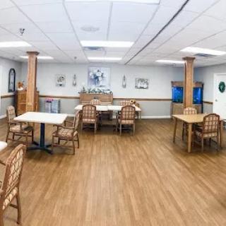 An empty dining area with wooden flooring, featuring tables and chairs arranged in a well-lit room.