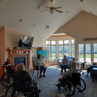 A group of seniors sitting in a communal living room, some in wheelchairs, engaged in a social gathering.