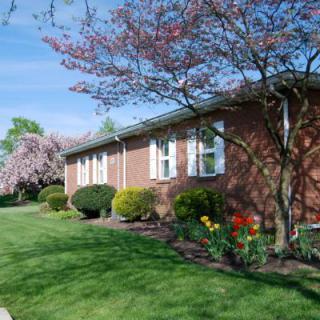 A brick house surrounded by green grass, blooming trees, and vibrant flowers in a landscaped garden.