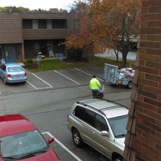 A worker in a bright yellow vest collects trash in a parking lot with autumn foliage.