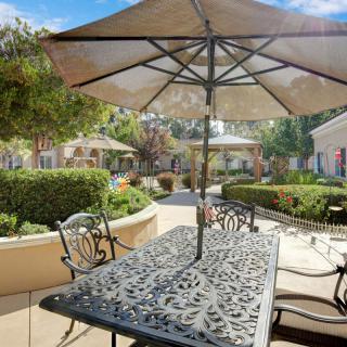 A sunny patio with a metal table and chairs under a large umbrella, surrounded by garden greenery.