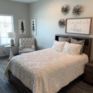 A modern and cozy bedroom with a neatly made bed, an upholstered chair, wall art, and a lampshade by the window.