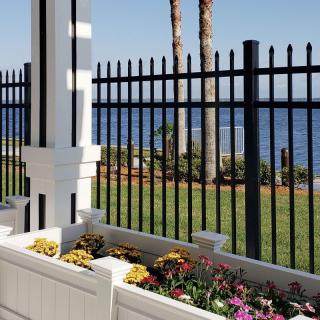 A garden with colorful flowers overlooks the ocean, framed by a black metal fence and palm trees.
